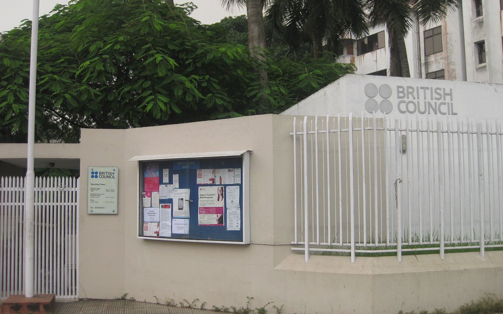 The entrance wall and notice board of the British Council office in Dhaka, with trees behind the white boundary fence