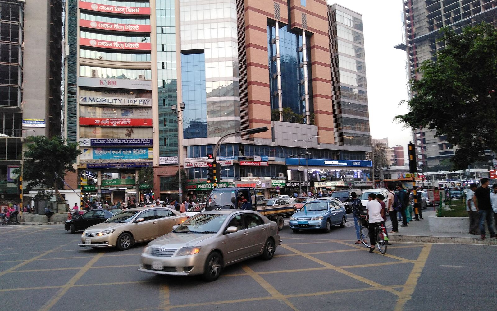 Traffic crossing Gulshan 2 circle in Dhaka in front of glass and brick office towers with shop signs at street level