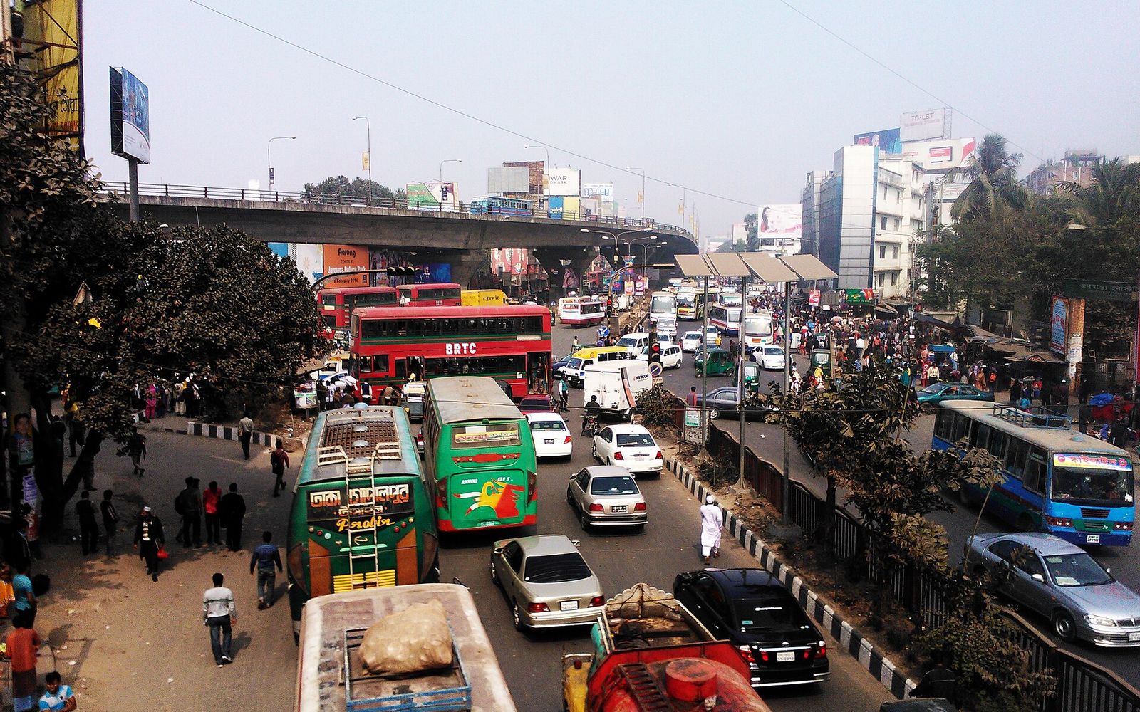 Buses and cars in heavy traffic under the Mohakhali flyover in Dhaka, with shops and office buildings along the road